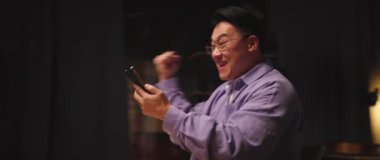 Merak ediyorum, gözlüklü Asyalı adam cep telefonu kullanıyor sosyal medyada sohbet ediyor gece evde mutlu dans ediyor. 360 derece. Kamera hareket ediyor