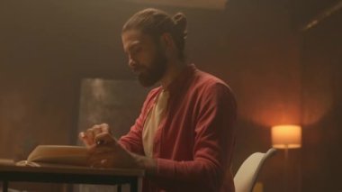 Odaklanmış genç erkek işçinin akşamları elinde belgelerle masasında oturduğu görüntüler. Sakallı, saç stili çarpıcı bir adam. Defterde yazıyor. Modern ofis. Arkaplanda renkli notlar olan şık bir duvar