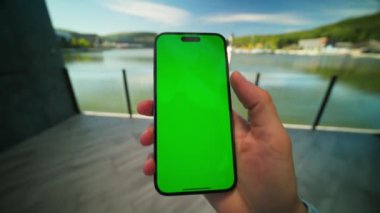Yeşil ekranlı akıllı telefonun yakın görüntüsü. Cep telefonu erkek elinde. Kromakey ekranlı akıllı telefon görüntüsü. Tanınmayan bir adam gölün güzel manzarasını çekmek için yaklaşıyor.