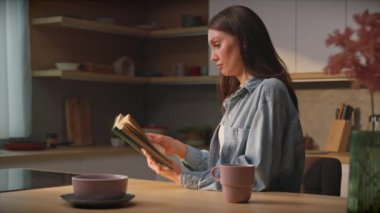 Kahverengi saçlı, beyaz kitabın mutfak masasında oturan genç bir kadının yan görüntüsü. Bayan okuyucu yalnız kalmanın tadını çıkarıyor. Günlük rutin. Modern daire