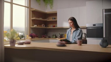 Modern mutfakta uzun saçlı, kot gömlek giymiş, kitap okuyan şık esmer bir kadının çekimleri. Rahatlama. Sıcaklık. İzin günü. Kapalı