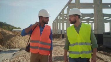 Beyaz kasklı, turuncu ve sarı yelekli Türk ve Kafkas mühendislerinin inşaat sahasında yürüdükleri görüntüler. İş günü. Jeodezi, endüstriyel kavram. Açık havada.