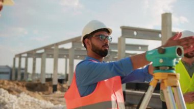 Açık havada birkaç mühendis bir inşaat projesi hakkında konuşuyor. Optik seviyeler kullanıyorlar. Güneş ışığı altında kask ve üniforma içinde durmak. İnşaat şirketi