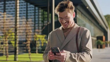 Sokakta gülümseyen bir telefonla yürüyen mutlu genç adam. Güneş ışığı var. İş adamı. Sosyal medya. Yavaş çekim