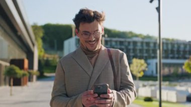Gözlüklü mutlu bir gencin sokakta gülümseyen bir telefonla yürümesi. Güneş ışığı var. İş adamı. Sosyal medya. Yavaş çekim