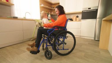 Babasıyla tekerlekli sandalyede oturan gururlu bir babanın fotoğrafı. Babası küçük bebeği besliyor. Ebeveynlik. Güzel görünümlü, çocuklu, gülümseyen beyaz bir adamın görüntüleri. İnsan hakları, eşitlik. Kişi: