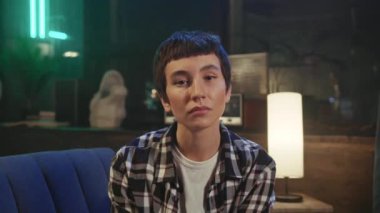 Ciddi şekilde odaklanmış, kendine güvenen, şık saç kesimli genç bir kadının portresi koltukta oturuyor ve akşamları rahat ofisin bulanık arka planında kameraya bakıyor. Yaratıcı kişi. Kapalı