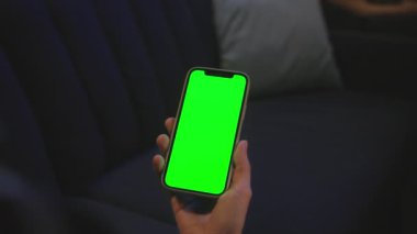 Tanımlanamayan bir kadının elinde akşam vakti yeşil ekranlı akıllı telefon görüntüsü. Ekose gömlekli bir kadın bir ekran alete dokunuyor, arka planda mavi kanepe kapalı.