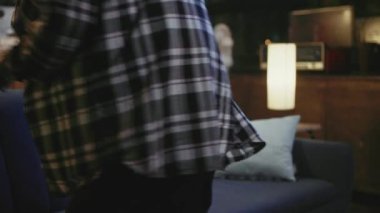 Modern oturma odasındaki kanepede otururken elinde dijital tabletle gülümseyen sevimli genç hippi kadın. Ekose gömlekli kız ekrandan aşağı bakıyor.