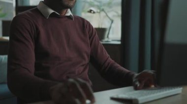 Tıraşsız Afrikalı Amerikalı bir adamın bilgisayarın önünde oturup dijital fare kullanarak klavyeye yazı yazması. Rahat bir ofisten çalışan serbest çalışan biri. Uzaktan çalışma konsepti. Kapalı