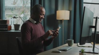 Rahatlamış gülümseyen Afrikalı adamın portresi akıllı telefonu elinde tutuyor, ekrana bakıyor, sohbet ediyor. Çalışanların işten izin alıp eğlendiği bir fotoğraf. Modern çatı katında akıllı telefon kullanan serbest çalışan.