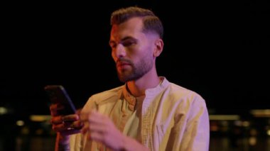 Ciddi bir genç adam gece vakti akıllı telefon kullanarak sosyal medyayı tarıyor. Açık havada. Kamera hareket ediyor