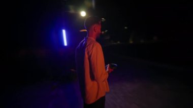 Kulaklık takan ve dışarıda müzik dinleyen genç bir adam tek başına eğleniyor. Gece kentinde duruyor