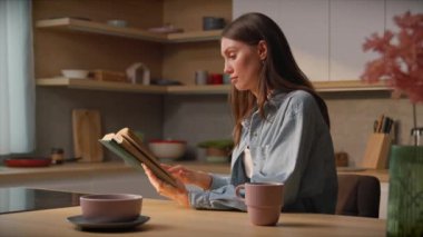 Güzel bir esmer, uzun saçlı, kot gömlekli, modern mutfakta kitap okuyan, mutfak masasında oturan. Bayan okuyucu sessizliğin tadını çıkarıyor. Günlük rutin. İçeride. Ev