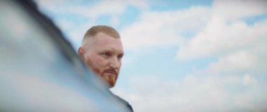 Dağlık ormanda arabadan inen şapkalı genç sakallı adamı kapatın. Arka planda mavi gökyüzü