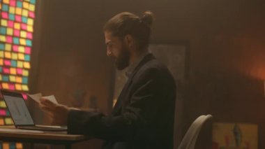 Sakallı genç iş adamı, masa başında oturup kağıtlarla çalışmayı düşünür. Gece ofisindeki bilgisayarı kullan. İşine odaklan. Akşam ışığı