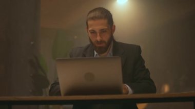 Çekici sakallı iş adamı masasında oturup çalışırken mutlu bir şekilde gülümsüyor. Gece ofisinde dizüstü bilgisayar kullan