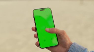 Sokakta elinde telefon krom anahtar yeşil ekran tutan genç adam. Akıllı telefonlu, tanınmayan beyaz bir adamın görüntüsü. Doğa. Yakın plan. Model çekim. Teknoloji, cihaz.