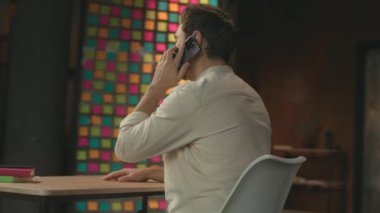 Masada oturup telefonla konuşan mutlu iş adamı akıllı telefondan gülümsüyor. Modern ofis. Sosyal medya. Kamera hareket ediyor. 360 Derece