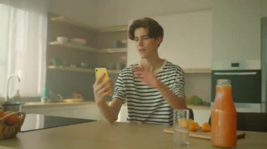 Beyaz erkek öğrencinin elinde akıllı telefonu tutarken ve video aramasıyla aradığı görüntüler. Genç yakışıklı adam kameraya bakıyor ve mutfakta el sallıyor. Kamera hareket ediyor. Gündüz vakti. Sıcak atmosfer