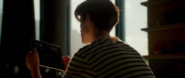 Dijital tableti elinde tutan ve alete bakan odaklanmış bir gencin görüntüsü. Uygulamalardaki mesajları kontrol eden adam. Beyaz tenli erkek öğrenci sıcak mutfakta ekrana dokunuyor. Gündüz vakti. Güneş ışığı