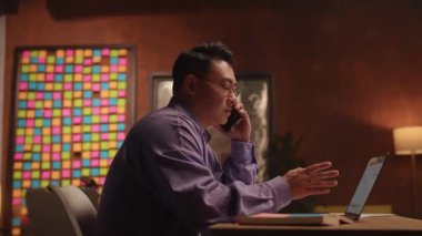 Gözlüklü Asyalı iş adamı dizüstü bilgisayar kullanıyor ve telefonda konuşuyor. Geceleri ekrana ciddi ciddi bak. Kağıt üzerinde çalışmak. 360 derece. Etrafta dolanıyorum.