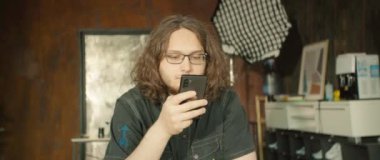 Odaklanmış genç bir adam şık, çağdaş bir stüdyo ortamında otururken akıllı telefonunu dikkatle kontrol eder.