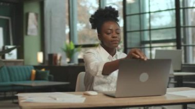 Modern ofiste, koltukta oturan Afro-Amerikan iş kadını dizüstü bilgisayar kullanarak gülümsüyor. Kamera hareket ediyor. 360 derece