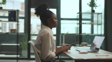 Sandalyede oturan Afro-Amerikalı iş kadını ofiste dizüstü bilgisayar ve telefon kullanıyor. Çok ciddiyim. Kamera hareket ediyor. 360 derece