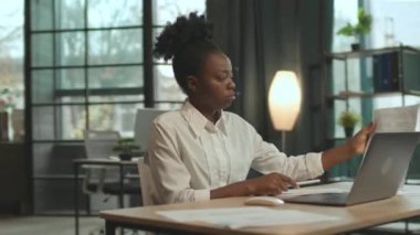 Ciddi bir Afro-Amerikan iş kadını koltuğunda oturuyor ve modern ofiste dizüstü bilgisayar kullanıyor. Kamera hareket ediyor. 360 derece