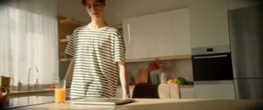 Çizgili tişörtlü beyaz erkek öğrenci masada oturuyor ve dizüstü bilgisayarla çalışmaya başlıyor. Genç adam evde online çalışıyor. Mutfakta rahat bir atmosfer var. Eğitim konsepti. Mesafe öğrenme. Kapalı