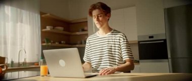 Mutfakta dizüstü bilgisayar kullanan genç bir erkeğin yatay görüntüsü. Evden çevrimiçi okurken öğrencilerin göz gezdirmesine odaklanmış. Uzak bir iş. İz sürme sahnesinde dolaşıyorum. İçeride. Gündüz vakti. Sinematik