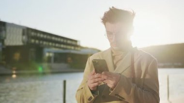 Gözlüklü ciddi bir genç adam gölün yakınındaki bir şehirde güneş ışığında cep telefonu kullanıyor. Açık havada duran kendinden emin genç iş adamları.