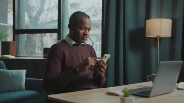 Akıllı telefona bakan, mesaj yazan ve sosyal medyayı karıştıran gülümseyen Afrikalı adamın yan görüntüleri. Evin rahat atmosferinde kahve molası sırasında neşeli bir serbest çalışanın portresi. Kapalı