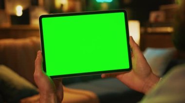 Adamın elinde yatay yeşil ekranla tablet bilgisayarını tıklatırken görüntüsü. Model yap. Arkaplanda kanepe