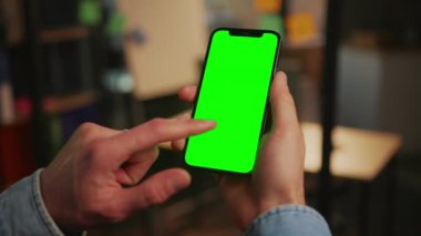 Sağ elin akıllı telefonu tuttuğu bir görüntü. Tanımlanamayan genç adam, günlük kot gömlek giyiyor yeşil ekrana vuruyor, alet kullanıyor. Krom anahtar kavramı. Dikey pozisyonda. Kapalı
