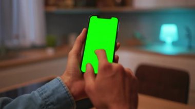 Modern mutfakta otururken, yeşil ekranlı akıllı telefon kullanan, tanınmayan genç adamların yakın çekimleri. Ekranı düzelt. İletişim konsepti. Krom anahtar. Kapalı