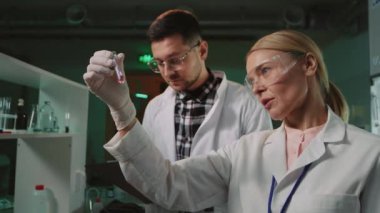 Koruyucu gözlüklü ciddi takım bilim adamları ışık laboratuarında elinde tüp tutan kadınla konuşuyor. Tıp, biyoteknoloji, kimya.