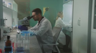 Laboratuvardaki mikroskoba bakan gözlüklü ve koruyucu eldivenli bir bilim adamı. Tıp, biyoteknoloji, kimya. Arka planda iş arkadaşım yürüyor.