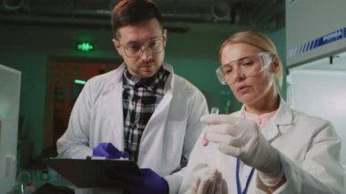 Koruyucu gözlüklü iki bilim adamı konuşan bir kadın ışık laboratuarında tüp tutuyor. Tıp, biyoteknoloji, kimya.