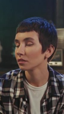 Kısa saçlı, kareli gömlekli, kendine güvenen genç beyaz bir kadının kanepede otururken ve akşam vakti masa lambasının bulanık arka planındaki kameraya bakarken yakın çekimi.