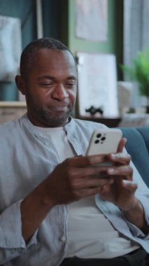 Rahat bir Afrikalı Amerikalı iş adamının sosyal medyada mesajları kontrol ederken dikey çekimi. Akıllı telefon kullanan gülümseyen bir adamın görüntüsü. İçeride. Bulanık arkaplan