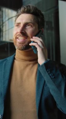 Şık ceketli, gülümseyen ve telefonda konuşan çekici bir genç adam iş merkezinde kendini mutlu hissediyor. Arka planda modern iç tasarım