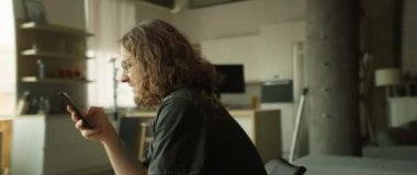 Kıvırcık saçlı bir insan, parlak ve modern bir yaşam alanında dinlenirken akıllı bir telefonla meşgul olur.