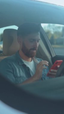 Genç bir adam arabada oturuyor, kot ceket giyiyor, akıllı telefonuna odaklanmış bir şekilde onunla etkileşimde bulunuyor.
