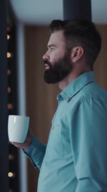 Sakallı bir adam pencerenin kenarında kahve yudumluyor, bir anlık içkiyle ve dışarıdaki manzarayla ısınan düşüncelerini yansıtıyor.