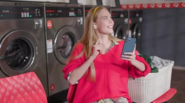 NEW YORK - 19 Aralık 2020: Güzel sarışın kadın çamaşır odasında oturmuş çamaşır yıkayan müzik dinleyen kulaklık ve telefon kullanıyor. Ev işi çamaşır makinesi kurutma makinesi içeride. Kapat.