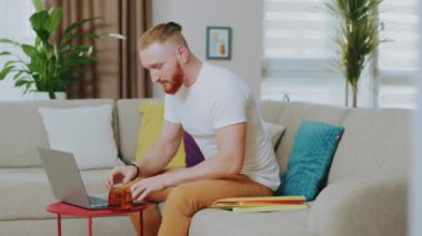 Çekici genç adam kanepede otururken dizüstü bilgisayar kullanıyor. Çay iç. Evde çalış. İnternet teknolojisi. Karantina zamanı. Kapatın. Yavaş çekim