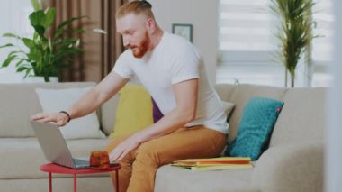 Mutlu genç adam kanepede otururken dizüstü bilgisayar kullanıyor. Çay iç. Evde çalış. İnternet teknolojisi. Karantina zamanı. Kapatın. Yavaş çekim