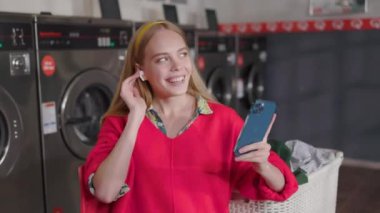 NEW YORK - 5 Kasım 2020: Gülümseyen neşeli genç sarışın kadın çamaşır odasında oturmuş çamaşır yıkayan müzik dinleyen kulaklık ve telefon kullanıyor. Ev işi çamaşır makinesi kurutma makinesi içeride.